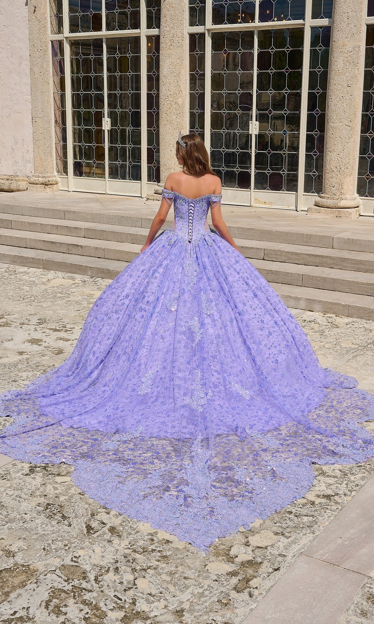 A woman wearing the Quinceanera Dress 54220 by Amarra, an off-shoulder floral glitter tulle ball gown with a long lace train, stands outdoors facing away before tall glass doors and stone columns.